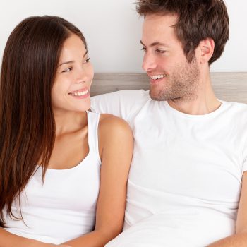Loving young couple sitting up in the comfort of their bed looking into each others eyes and smiling happily. Interracial couple, Asian woman, Caucasian man.